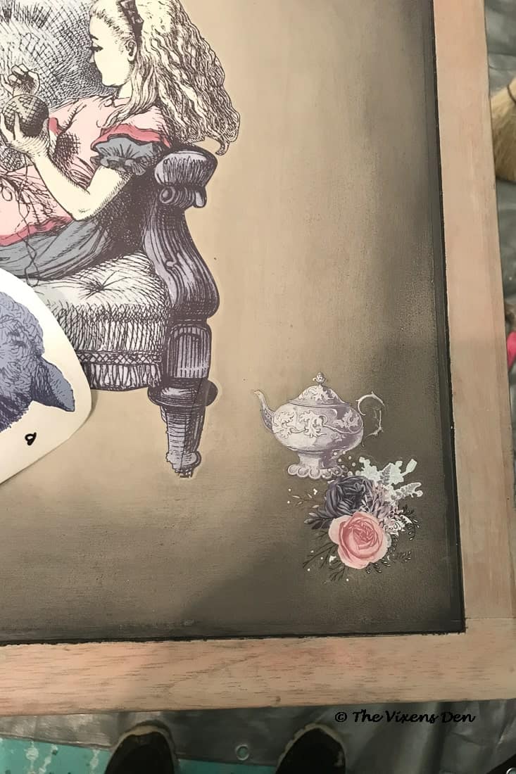 closeup of a large Alice in Wonderland transfer with smaller layered teapot and flower transfers on a cherry side table top painted in Dixie Belle French Linen and blended with Caviar along the edges