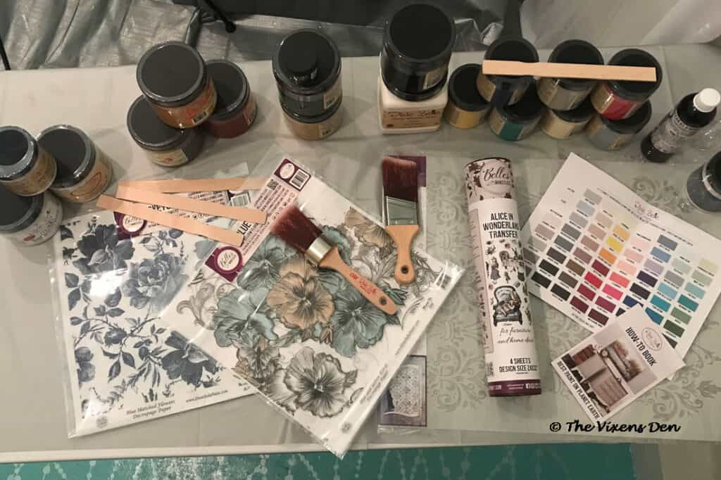 Dixie Belle painting supplies: stacks of paint, stir sticks, brushes, rice paper, a tube of transfers, paint swatchsheey and a How To Book