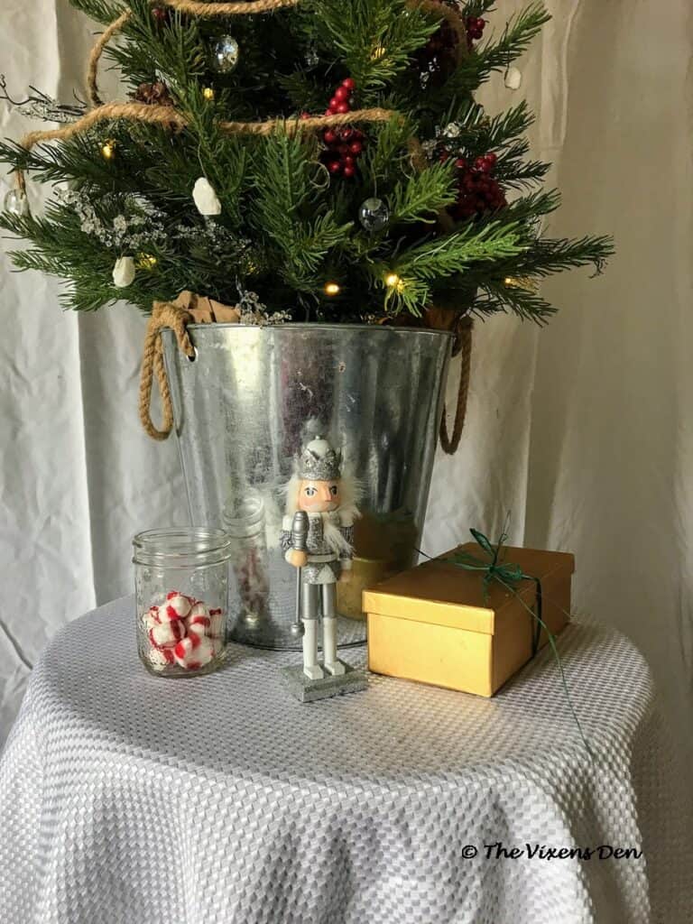 close up of mini Christmas tree base in metal trash can with a jar of peppermints, a silver nutcracker and a gold present