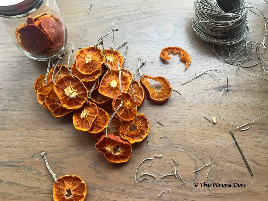 a pile of dried orange slices strung on hemp cord loops with a round of hemp cord, a large needle, and a jar of dried orange ends