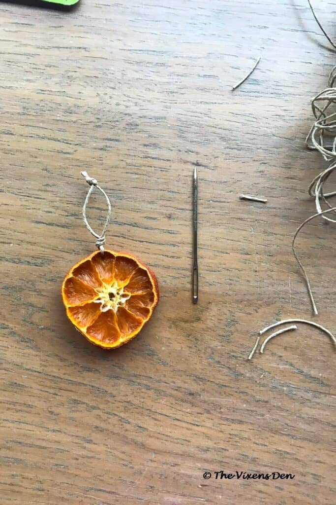 a dried orange slice with both ends of hemp cord knotted and a large needle laying next to it.