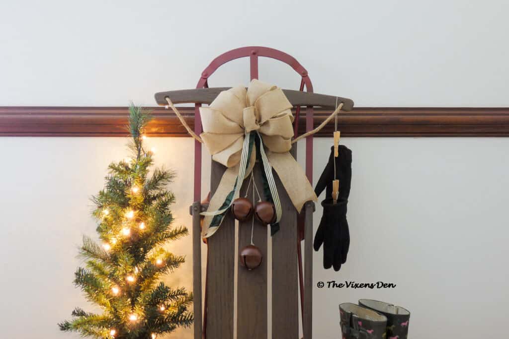 closeup of vintage sled staged with burlap bow, bells, boots, mini Christmas tree, and black gloves