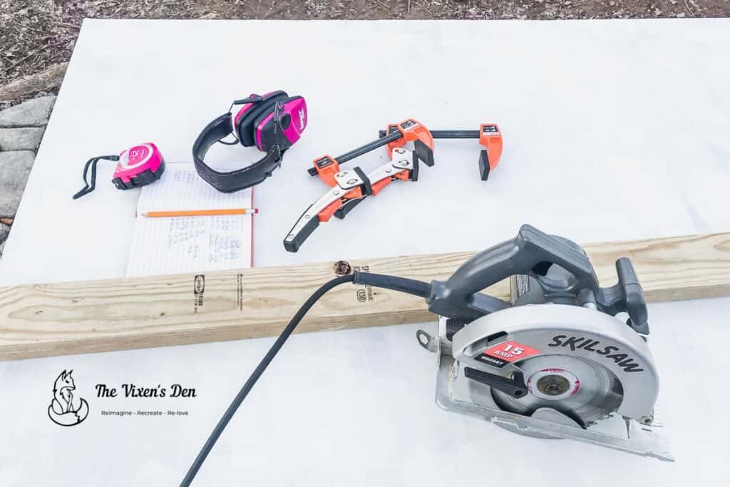 circular saw, 2x4, clamps, earmuffs, tape measure, notebook, on a sheet of faux brick paneling-min