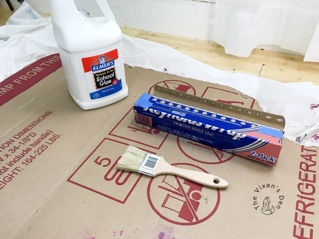 Elmers glue, aluminum foil and a paintbrush sitting on cardboard