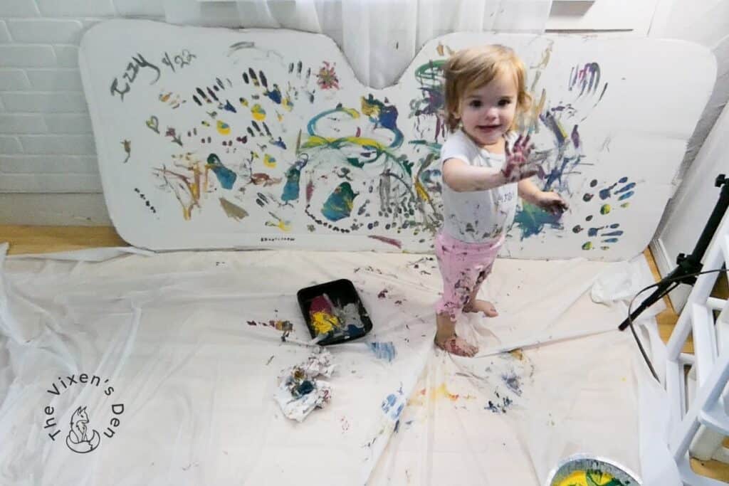 Toddler waving goodbye with a pan of paint and a painted sunshade in the background
