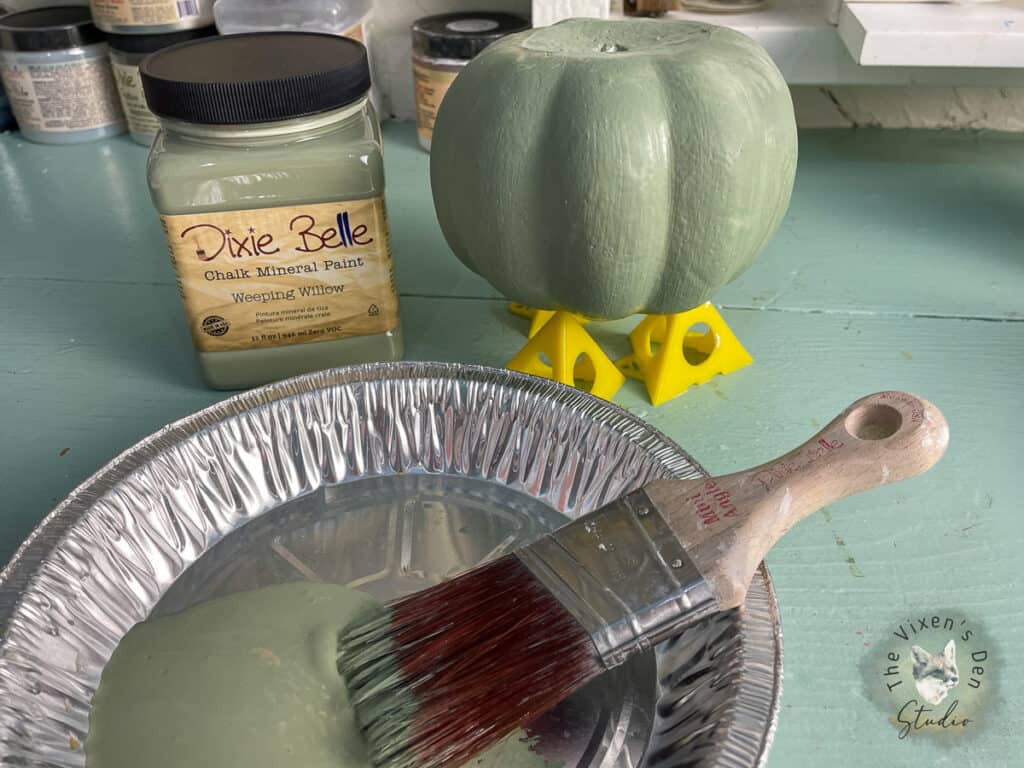 A green pumpkin on a table next to a brush and paint.