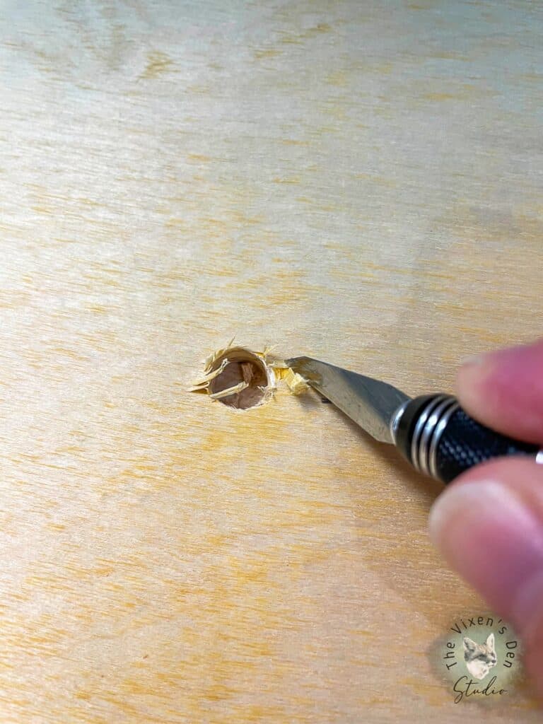 A person is using a craft knife to trim around a hole in a piece of wood.