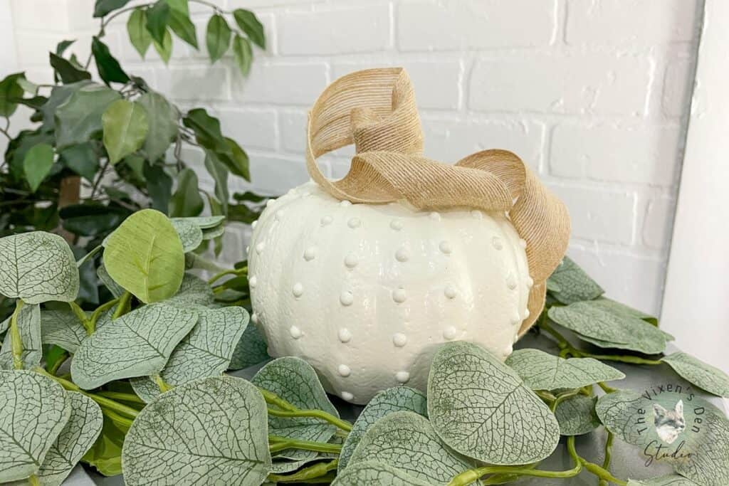 A white pumpkin sitting on a table with eucalyptus leaves.