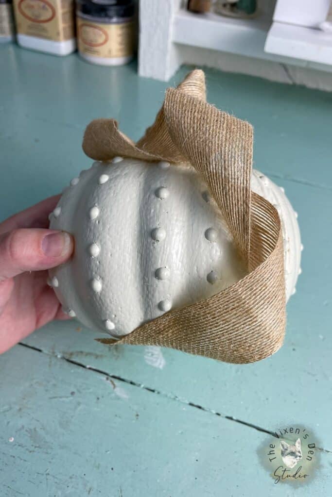 A hand holding a white pumpkin with a burlap ribbon.