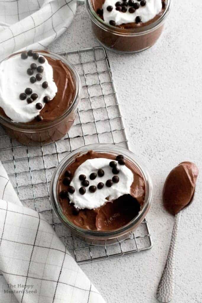 Three chocolate puddings with whipped cream and chocolate chips on a cooling rack.