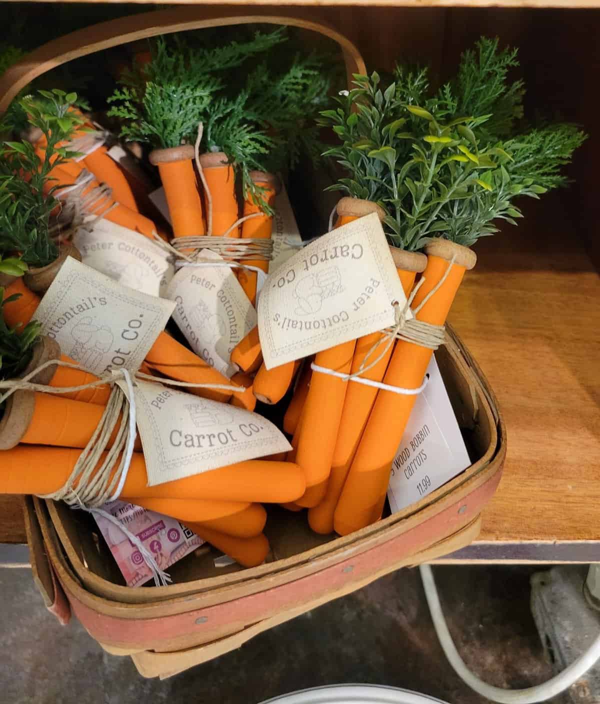 A basket filled with artificial carrot decorations made of fabric, arranged to resemble a bunch of harvested carrots, complete with faux greenery to mimic the tops.