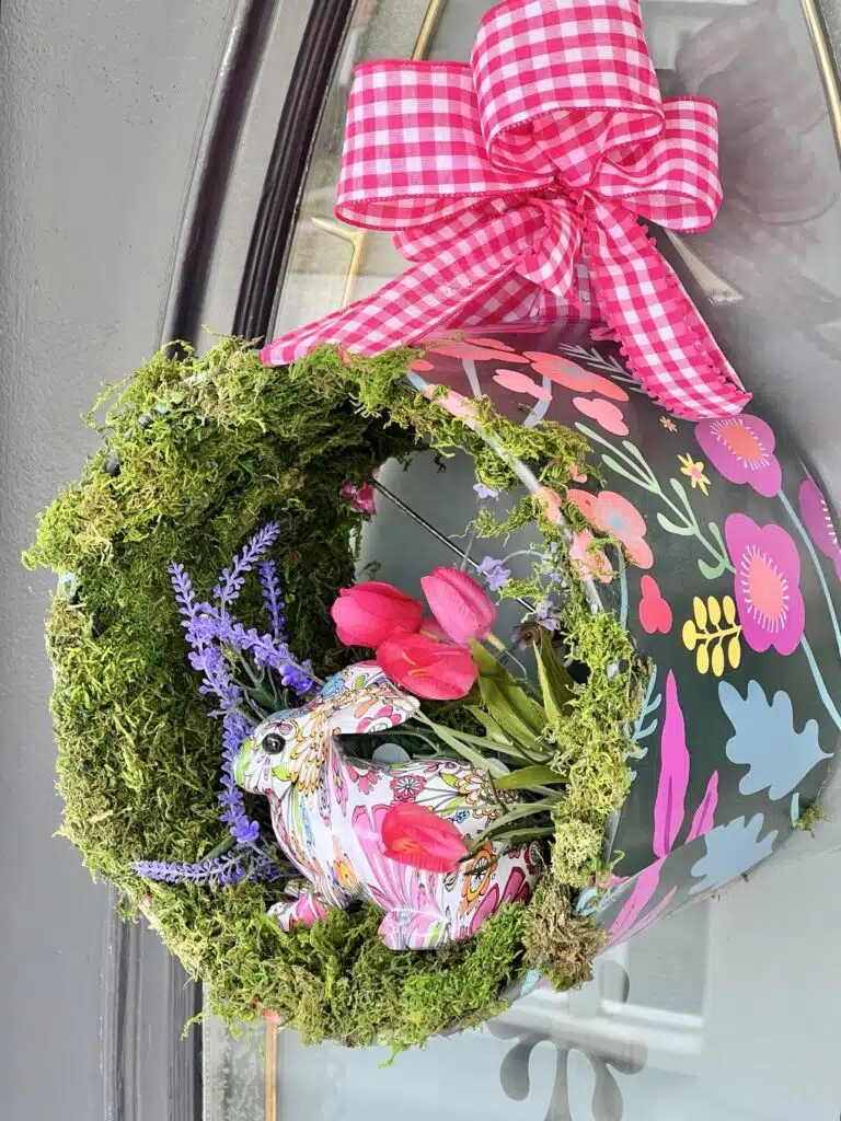 A decorative spring wreath with moss, faux lavender, pink flowers, and a patterned easter egg, adorned with a pink gingham ribbon.