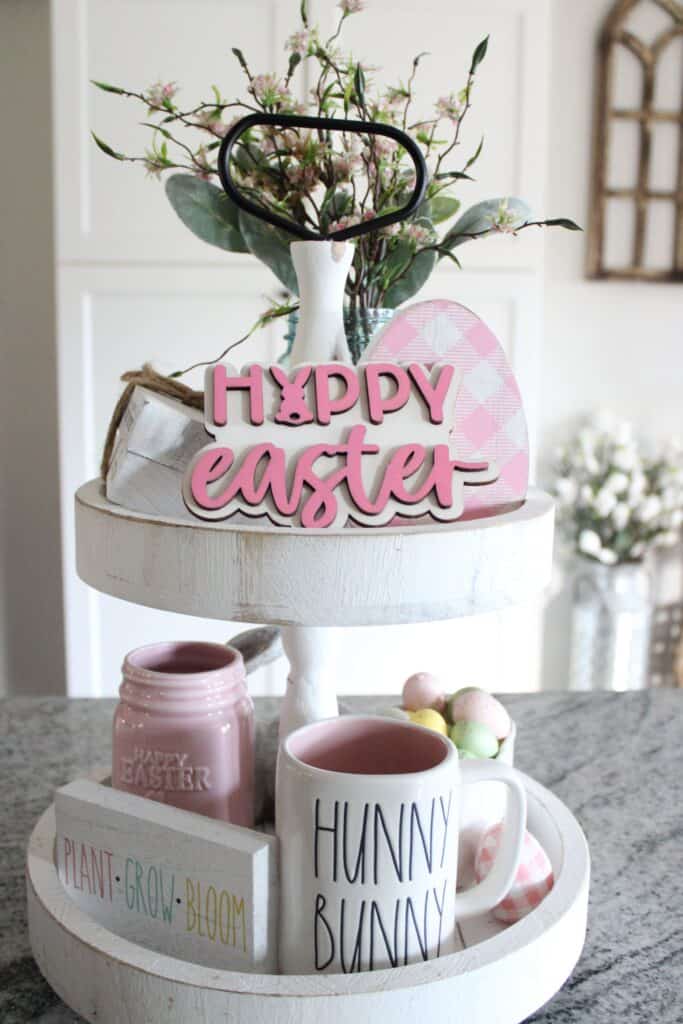 A two-tiered easter-themed decorative tray containing a floral arrangement, festive signs, and mugs.