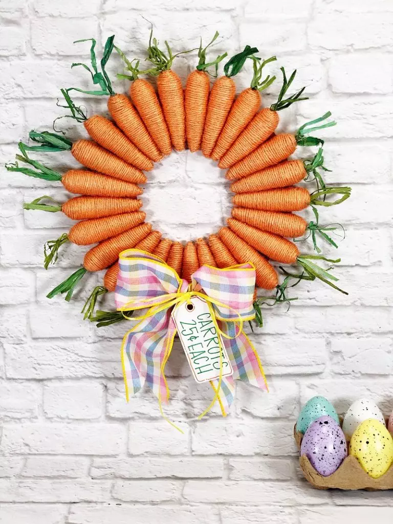 Decorative carrot-themed wreath with a bow and "happy easter" tag, displayed on a white brick wall next to a trio of speckled easter eggs.
