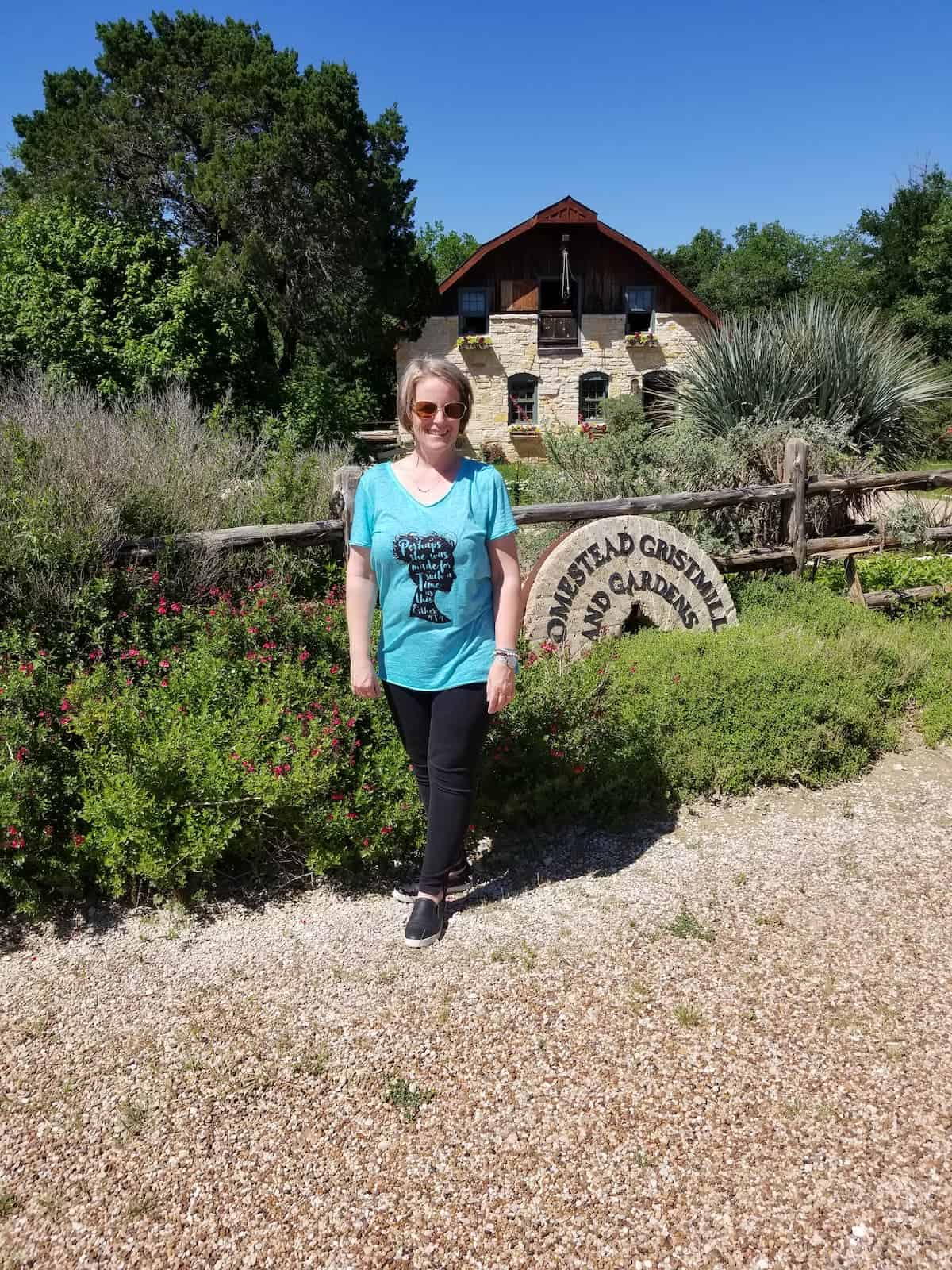 Woman standing in front of a rustic building with a sign reading "wildseed farms" surrounded by greenery and flowers.