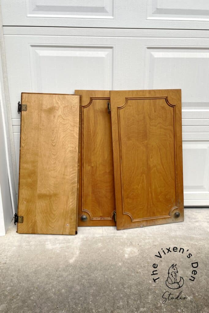 Two wooden cabinet doors leaning against a garage door, equipped with hinges and knobs.
