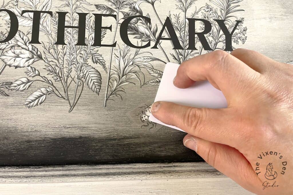 A hand holding an eraser removing a smudge on a wooden surface with botanical illustrations and the word "apothecary".