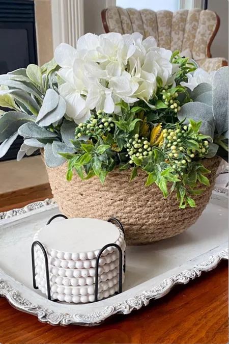 A decorative artificial flower arrangement in a woven basket placed on an elegant tray with a small patterned container beside it.