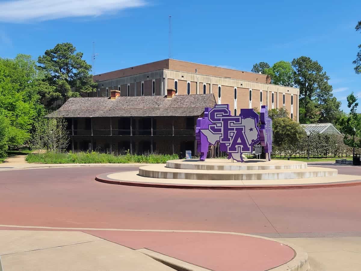 A university campus featuring a lush green lawn, a large stage with a purple stephen f. austin state university logo, and a multi-storied academic building in the background under a clear blue sky.