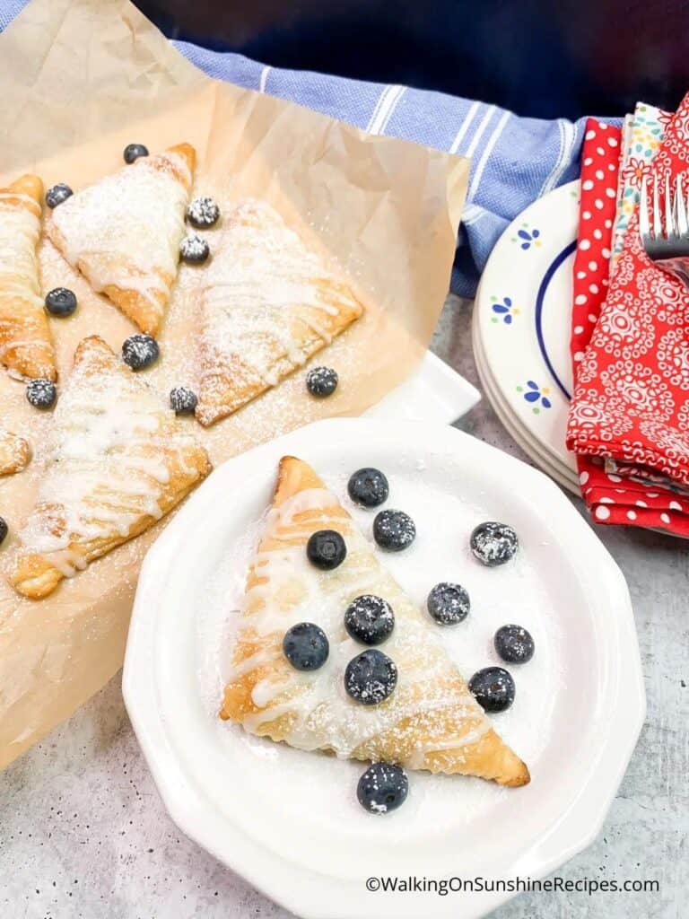 Powdered sugar-dusted pastries served with fresh blueberries on a white plate, with additional pastries in the background.
