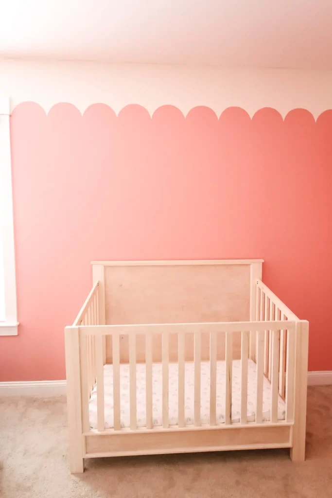 A natural wood crib in a nursery with pink walls and scalloped paint detail.