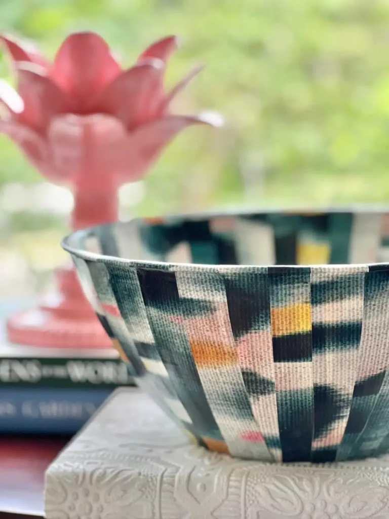 A patterned ceramic bowl in the foreground with a blurred pink plant in the background, set on a table with books.