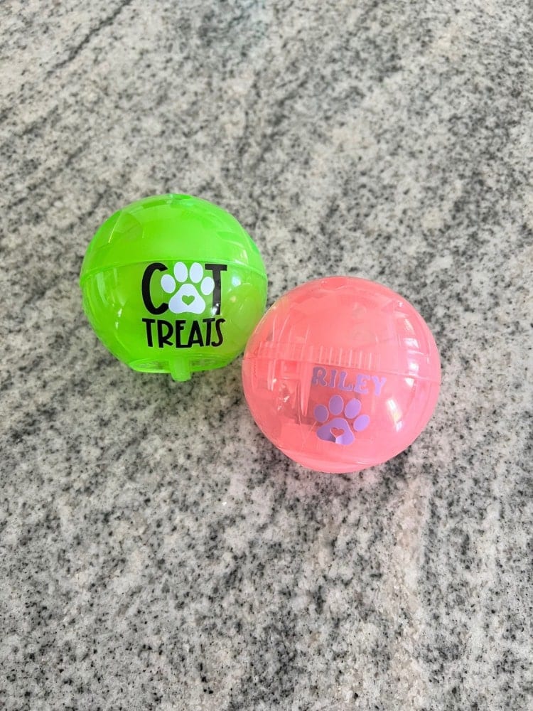 Two personalized dog treat balls, one green and one pink, with the names "cool treats" and "riley" on them, placed on a speckled gray surface.