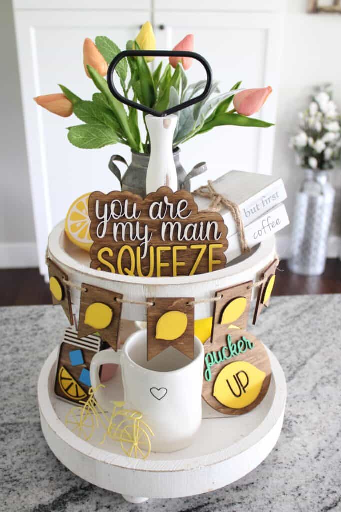 A decorative tiered tray featuring various lemon-themed ornaments, signs reading "you are my main squeeze" and "but first, coffee", and a bicycle motif.
