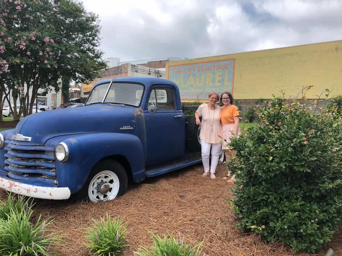 Two people standing beside a vintage blue truck with a colorful "the city beautiful laurel" mural in the background.