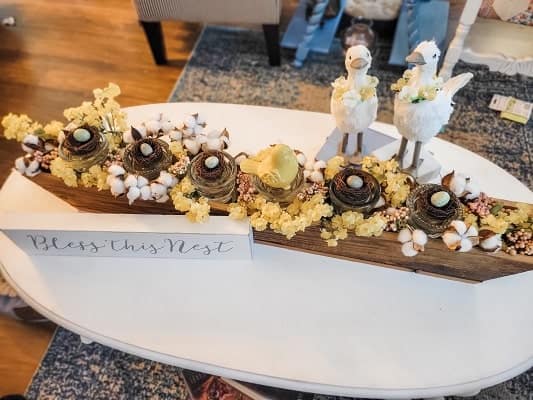 A decorative centerpiece featuring bird figures standing atop nests accented with flowers on a wooden board with the phrase "bless this nest" displayed in front.