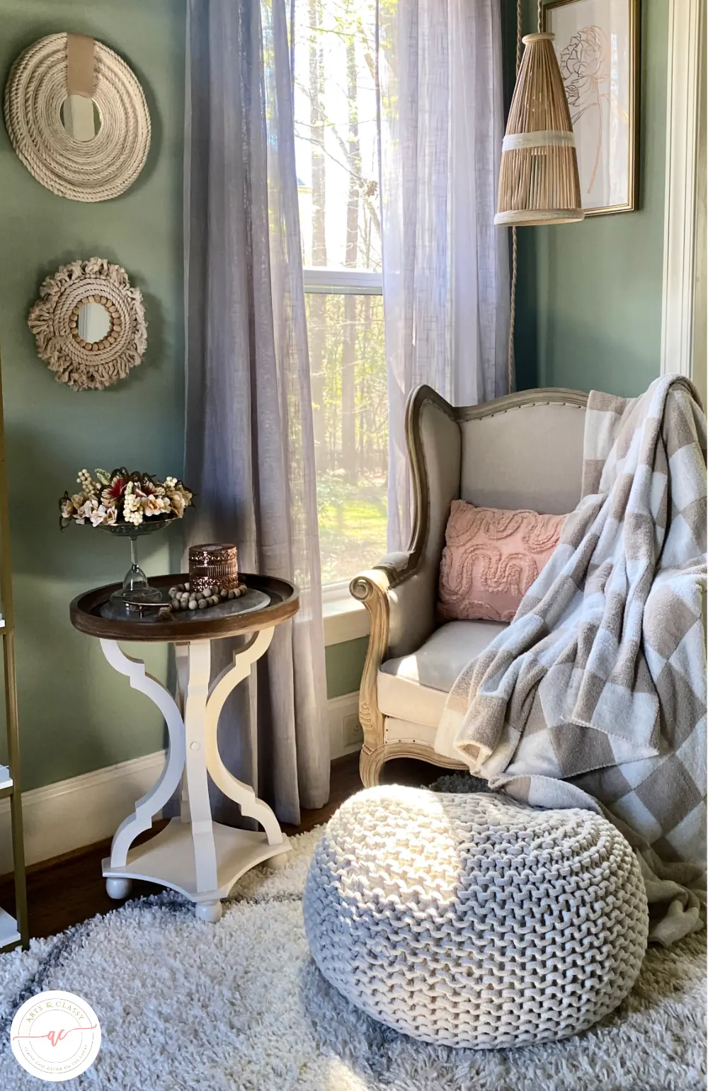 A cozy corner with a gray armchair, white side table, and knit pouf, set against teal walls and flowing curtains, illuminated by natural light.