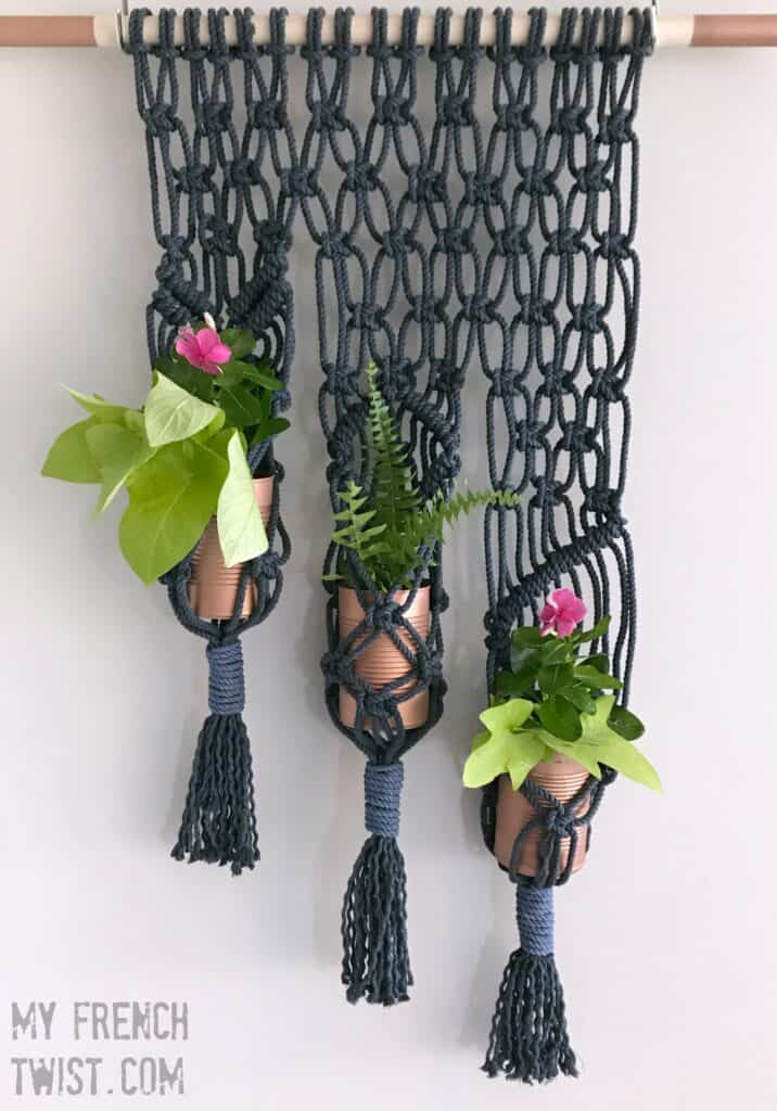 Three macramé plant hangers with copper planters, containing green plants and pink flowers, hung against a white wall.