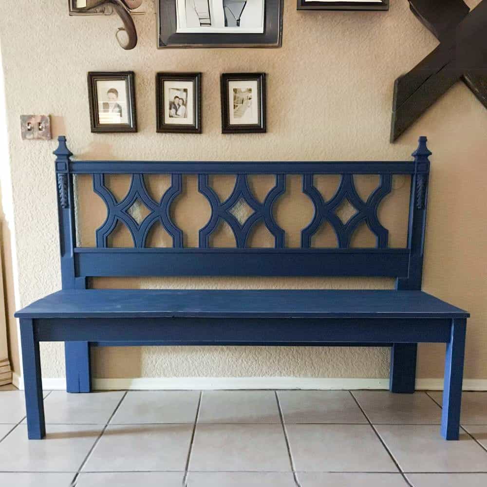 A blue wooden bench with a high backrest featuring ornate cut-out designs, positioned against a wall with family photos and a wall clock.