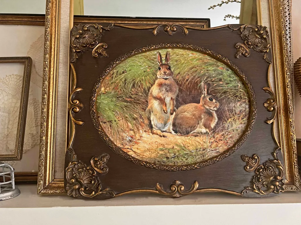 An ornate, oval-framed painting of two rabbits sitting in a grassy field, displayed on a shelf among other framed items.