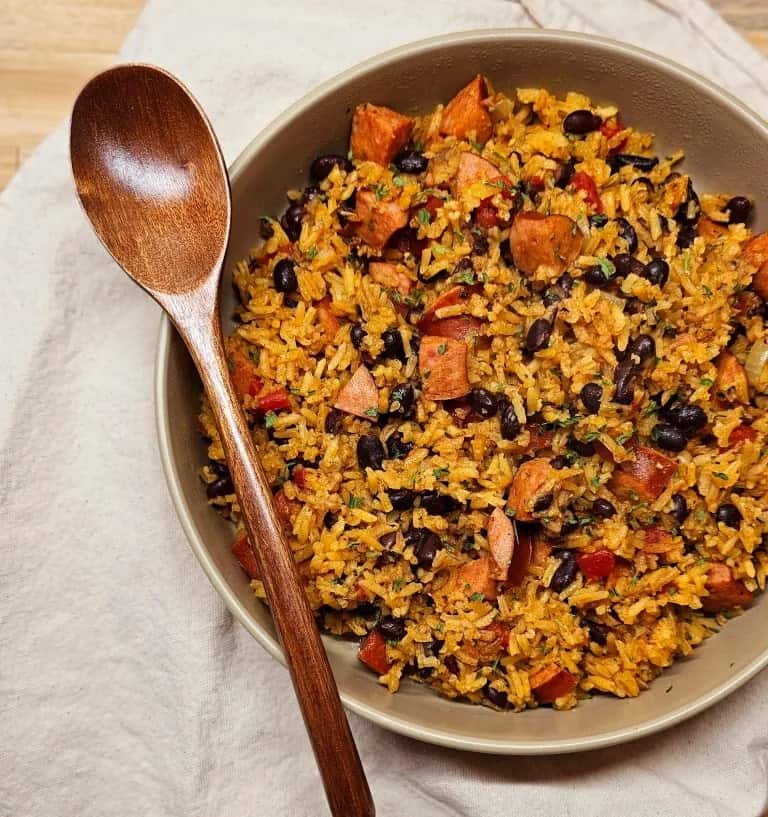 Bowl of seasoned rice mixed with black beans, diced vegetables, and sausage, accompanied by a wooden spoon on a white cloth.