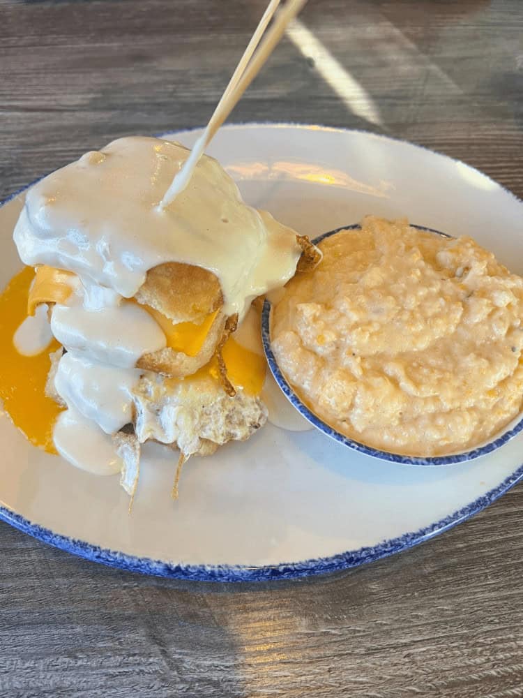 A plate with a biscuit sandwich topped with melted cheese and cream, and a side bowl of creamy grits. A toothpick is being placed into the sandwich.