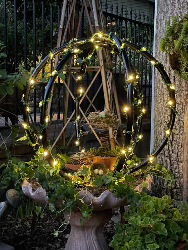 A garden scene featuring a spherical metal structure adorned with string lights, placed above a plant-filled fountain base amidst green foliage.