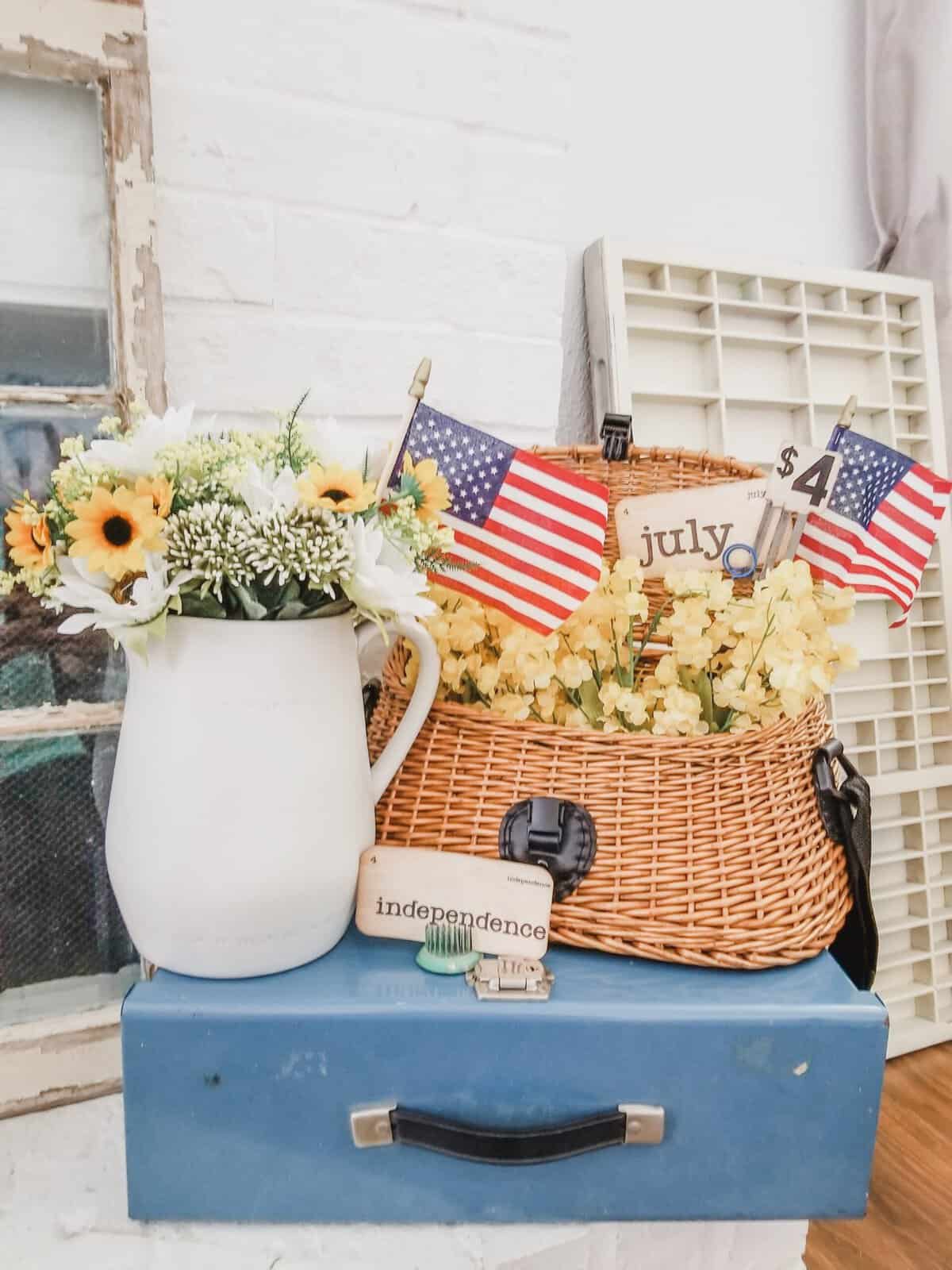 A blue table holds a white pitcher with flowers, a basket with yellow flowers, two small American flags, and wooden blocks spelling "independence" and "life." A sign reads "July 4.