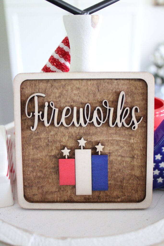 A wooden sign with the word "Fireworks" and three rectangular shapes with stars on top, colored red, white, and blue, against a rustic wooden background.