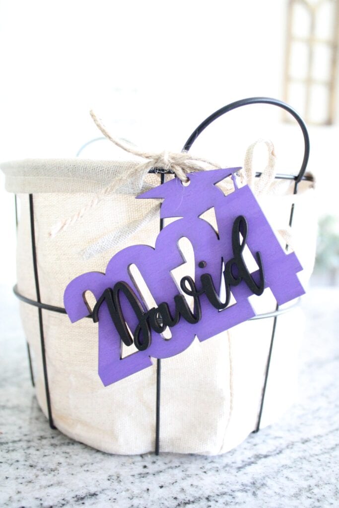 A striped fabric basket with a handle, adorned with a purple tag labeled "maria" in cursive script, placed on a kitchen counter.