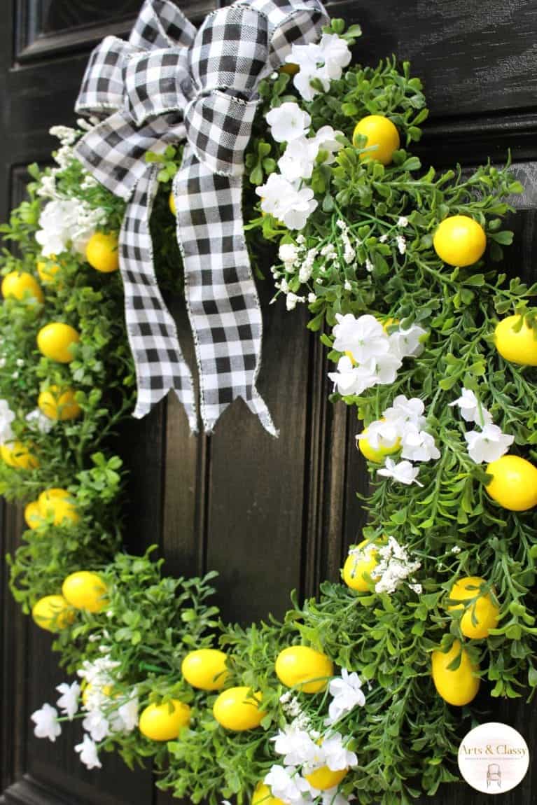 A decorative wreath with white flowers and yellow lemons, adorned with a black and white checkered ribbon, hanging on a dark wooden door.