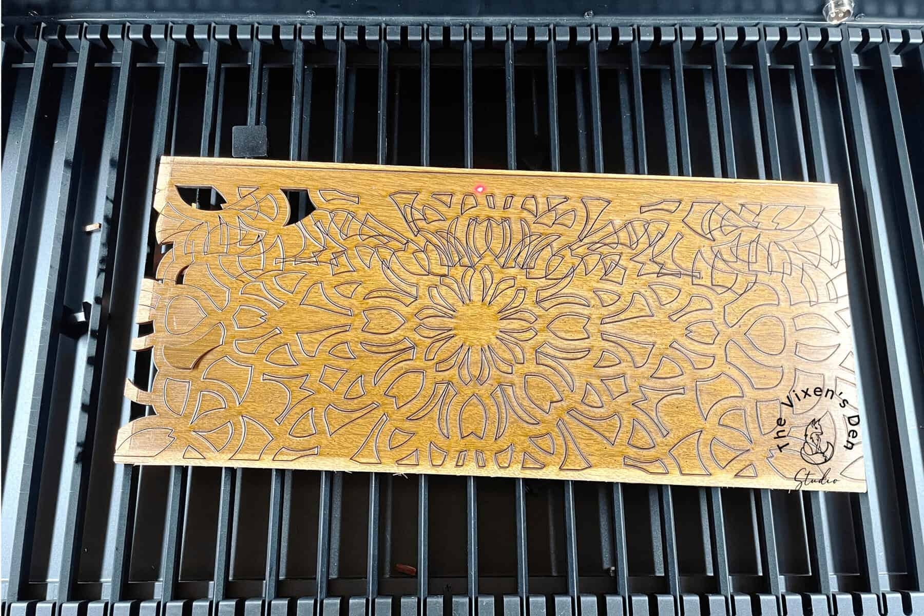 A rectangular wooden cutting board with intricate floral carvings sits on a black grill. One corner is broken off. The year "2021" and a signature are engraved on the bottom right corner.