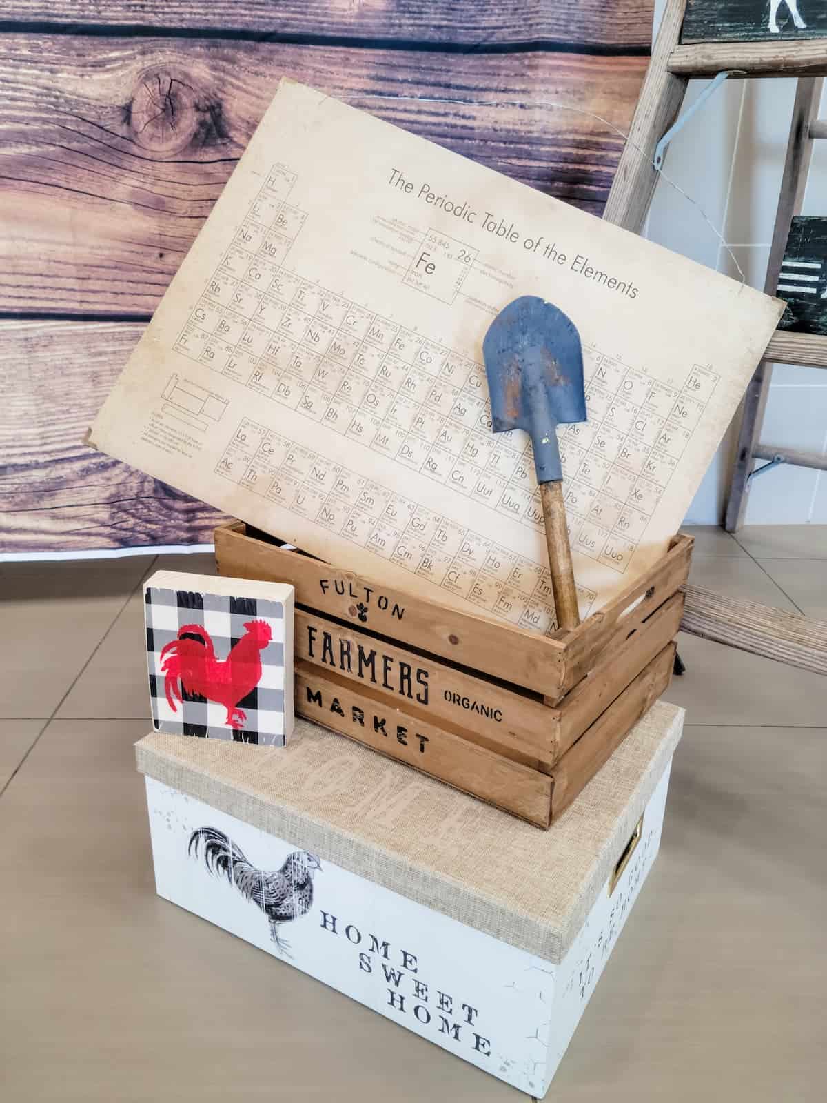 A decorative display with a shovel, periodic table of elements, wooden crate labeled "Farmers Market," a rooster sign, and a white "Home Sweet Home" box.