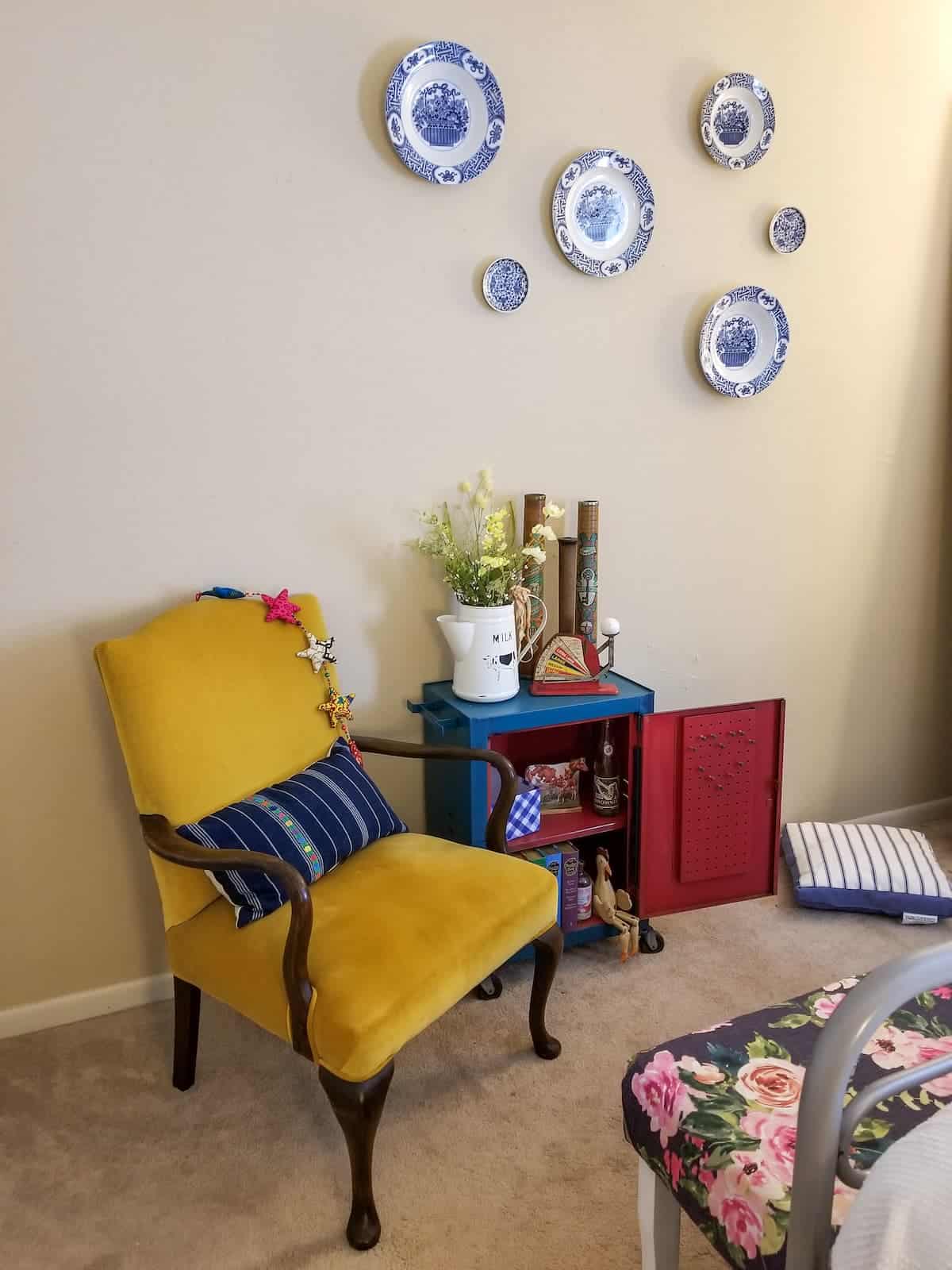 A cozy reading nook with a mustard yellow armchair, surrounded by decorative blue and white plates on the wall and a small side table with books and plants.