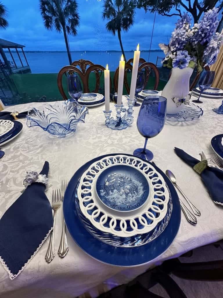 Elegant dinner table setup by the sea, featuring blue and white china, lit candles, and a floral centerpiece at dusk.