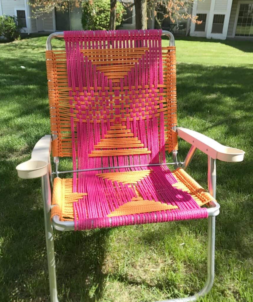 A lawn chair with a metal frame and woven pink and orange fabric positioned on a grassy lawn in front of a building.