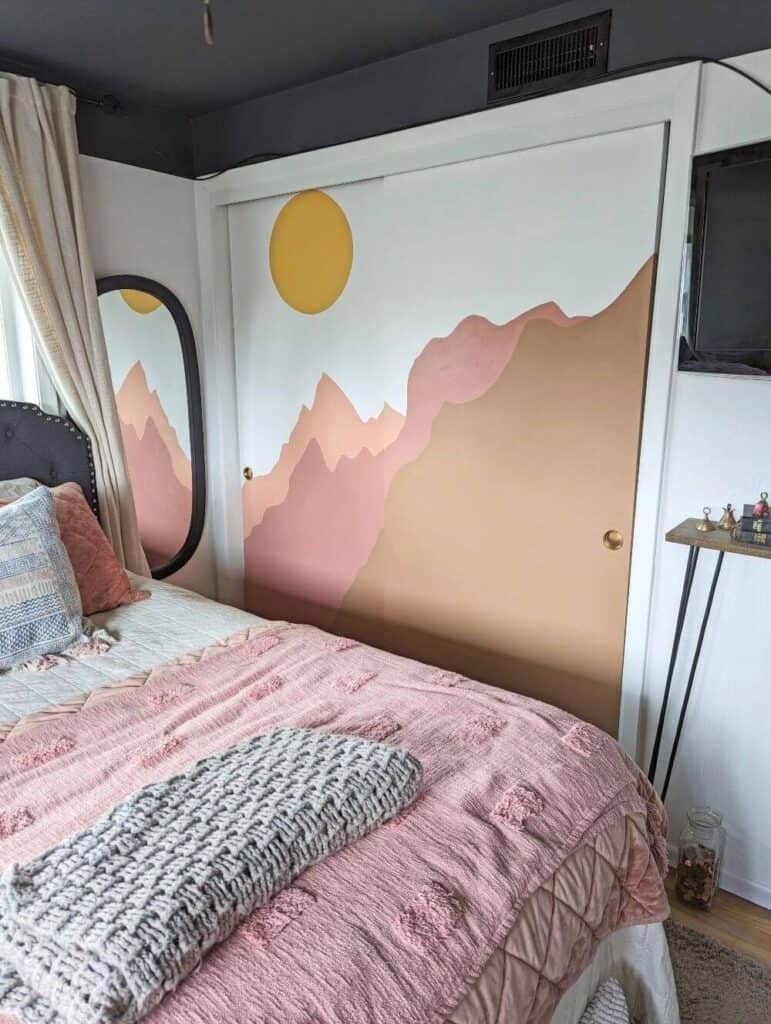 A bedroom features a bed with pink and beige bedding, a crochet blanket, and a mural of mountains with a sun on the closet doors. A mirror and a mounted TV are also visible.