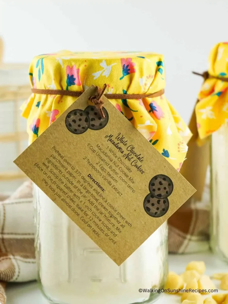 A jar of white chocolate hot cocoa mix with a yellow fabric lid cover and a label with a cookie design, highlighting ingredients and directions.