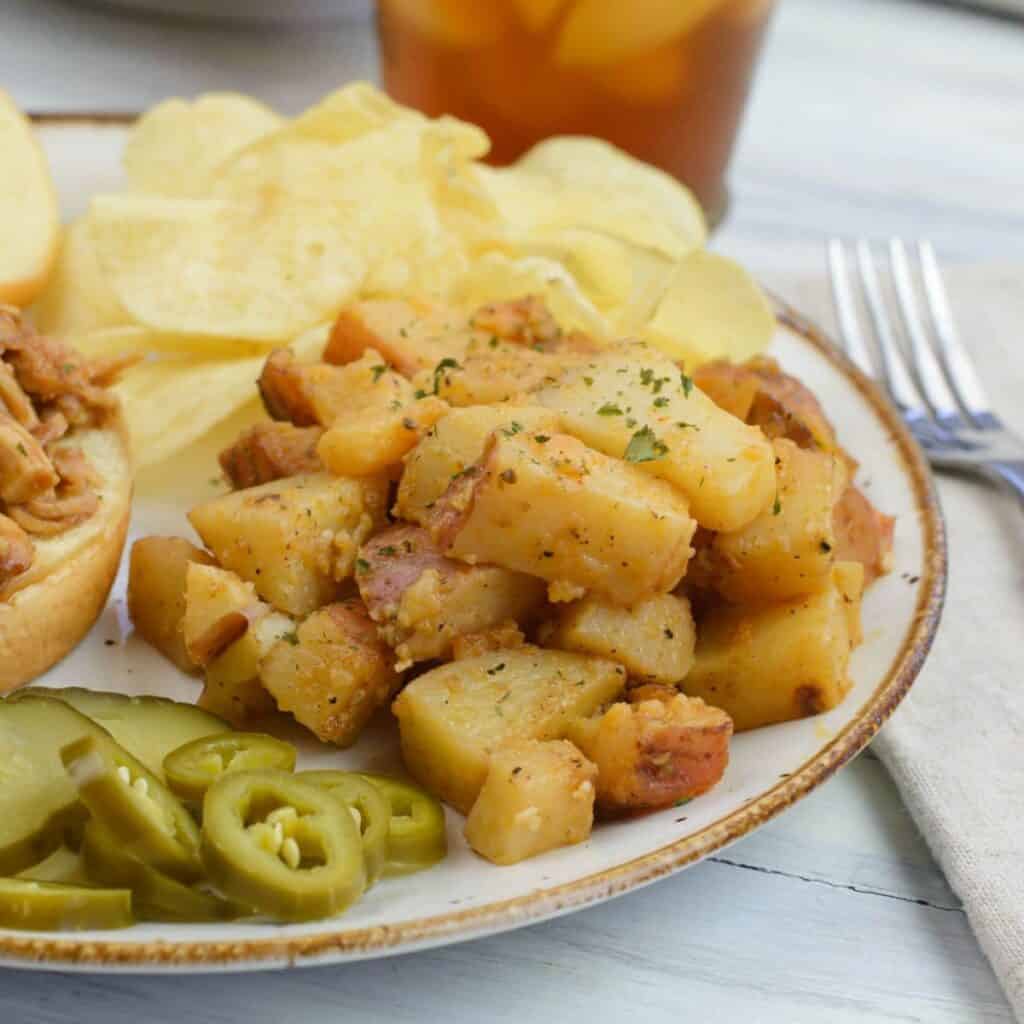 A plate with seasoned potato cubes, sliced pickles, jalapeño slices, potato chips, and part of a sandwich bun, accompanied by a fork and a glass of iced tea in the background.