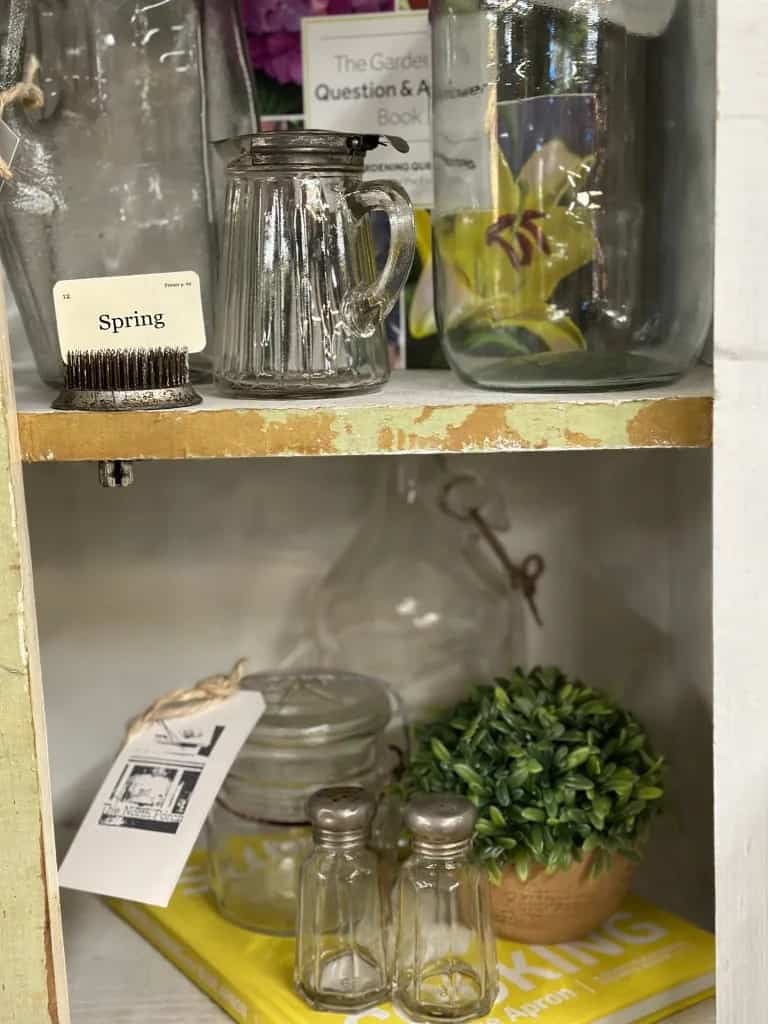 Various glass jars and containers arranged on wooden shelves, with decorative items including a small plant, a book, and a vintage brush labeled "spring.